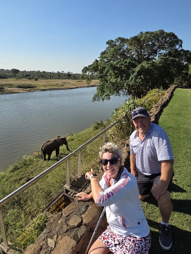 Two guests at a rail above a river, an elephant on the far bank below them
