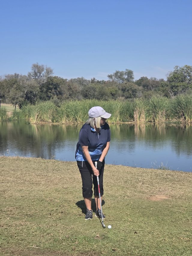 A golfer addressing the ball on mown grass beside still water, reeds and bush on the far bank