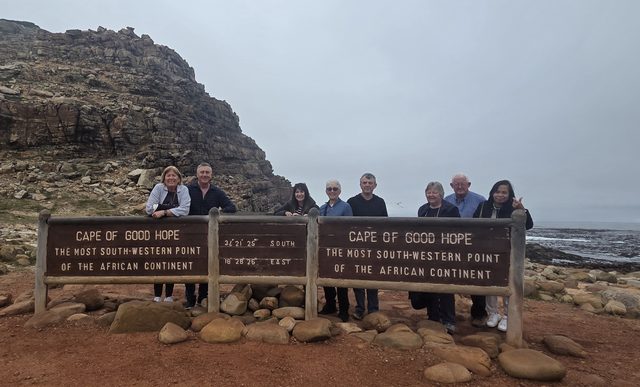 Nine guests behind the Cape of Good Hope sign, the headland rising behind