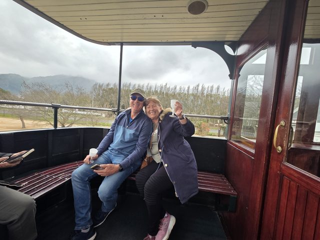 Two guests on the open deck of a wine-tram carriage, mountains and vines beyond the rail