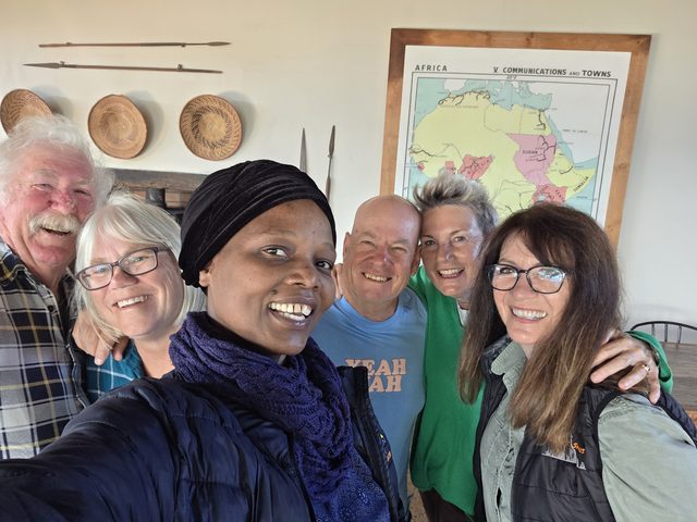 Six guests together indoors, a framed antique map of Africa and woven baskets on the wall behind them