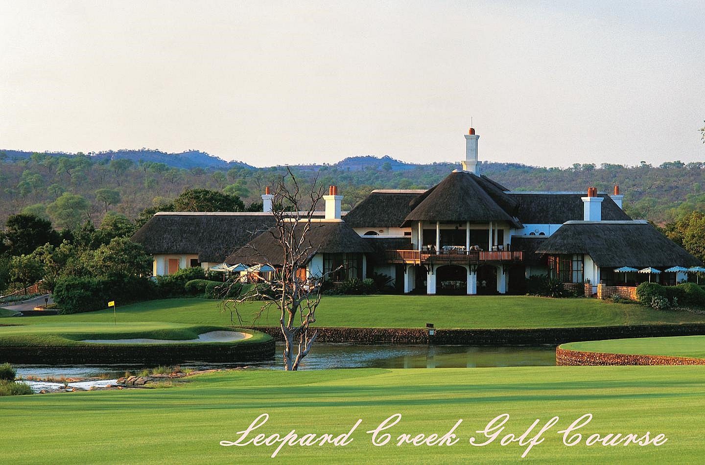 The thatched clubhouse at Leopard Creek seen across a pond, its white gables and chimneys above a covered terrace, with a bare tree on the near bank, a bunker and flag to the left, and bushveld hills behind. The fairway in the foreground carries the course's own script credit, Leopard Creek Golf Course