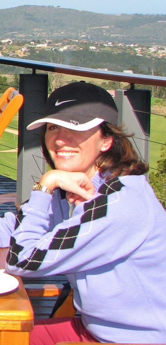 Sandy at a golf course, in a cap and sweater, leaning on a table with the fairway and hills behind her